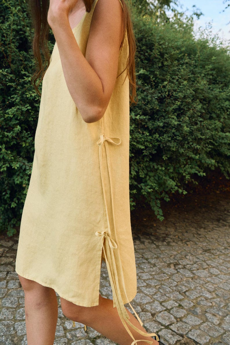 May include: A sleeveless, pale yellow linen dress with side ties. The dress has a simple, flowing design and a knee-length hemline. The side ties add a decorative element. The dress is made of a lightweight fabric, perfect for warm weather.