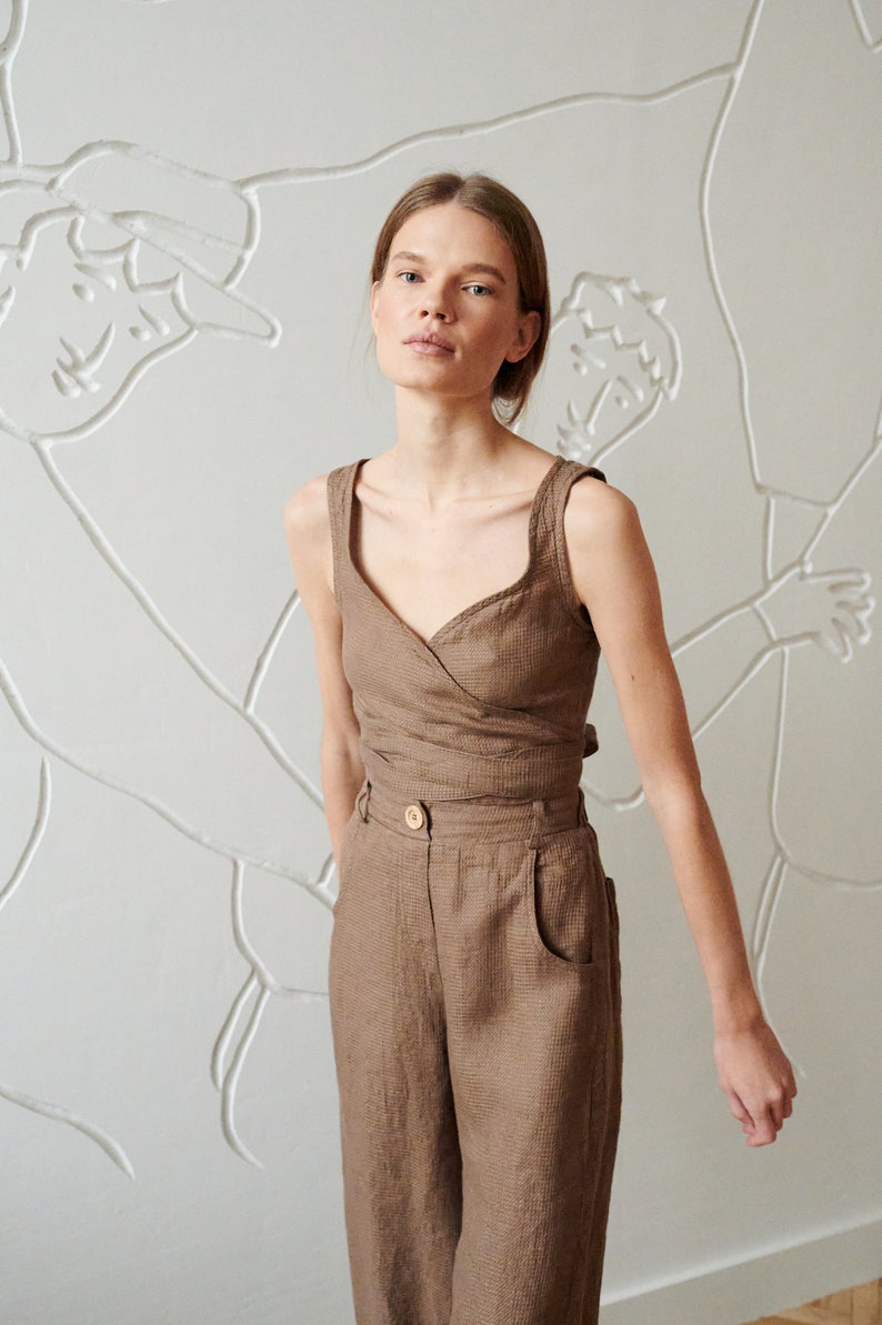 May include: A woman wearing a brown linen wrap top and matching wide-leg pants. The top has a V-neckline and a button closure at the waist. The pants have a high waist and pockets on the sides.