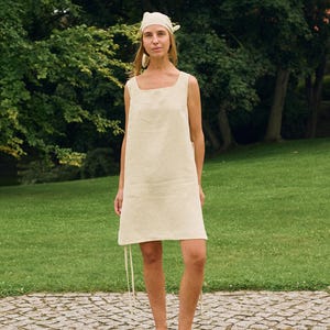 May include: A cream-colored linen dress with a square neckline and a matching headscarf. The dress has a simple, sleeveless design and falls to the mid-thigh. The model is wearing black leather slip-on shoes. The background is a green park.