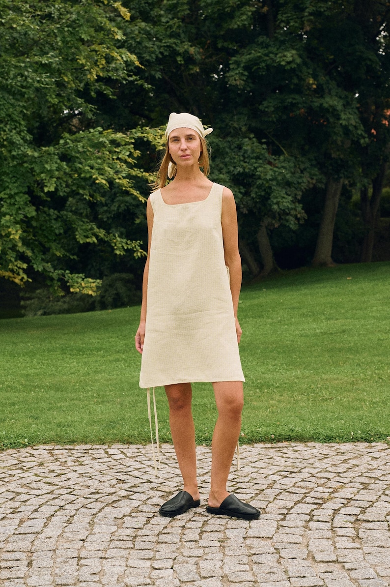 May include: A cream-colored linen dress with a square neckline and a matching headscarf. The dress has a simple, sleeveless design and falls to the mid-thigh. The model is wearing black leather slip-on shoes. The background is a green park.