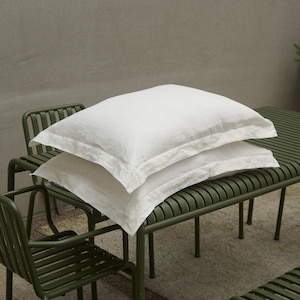 May include: Two white linen pillowcases are stacked on a green metal bench. The bench and matching chairs have a slatted design. The pillows have a soft, slightly wrinkled texture, suggesting a comfortable, relaxed aesthetic. The scene is set outdoors.