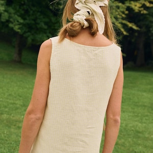May include: A sleeveless, light yellow dress with a square neckline. The person is wearing a matching headscarf tied in a knot. The background is a green, grassy area with trees.