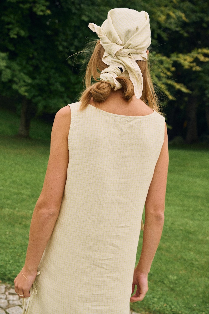 May include: A sleeveless, light yellow dress with a square neckline. The person is wearing a matching headscarf tied in a knot. The background is a green, grassy area with trees.