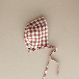 May include: A red and white gingham bonnet with a tie. The bonnet is made of a soft, lightweight fabric.