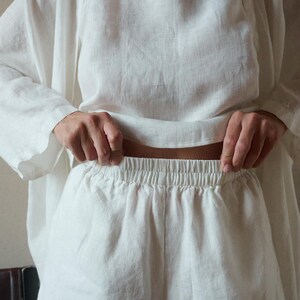 May include: A white linen outfit consisting of a long-sleeved top and shorts. The top has a round neckline and loose fit. The shorts have an elastic waistband. The fabric appears to be lightweight and breathable, suitable for warm weather.
