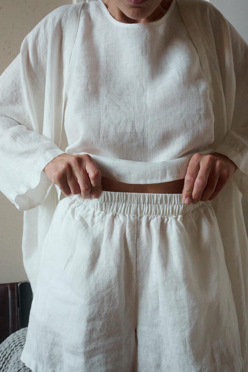 May include: A white linen outfit consisting of a long-sleeved top and shorts. The top has a round neckline and loose fit. The shorts have an elastic waistband. The fabric appears to be lightweight and breathable, suitable for warm weather.