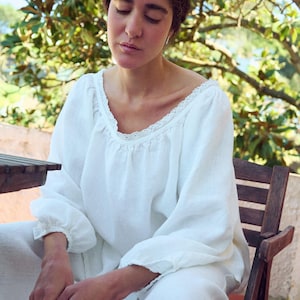 May include: A person wearing a white linen top and pants. The top has a scoop neckline with lace trim and long sleeves. The person is seated on a wooden chair, with a natural background of trees and foliage.