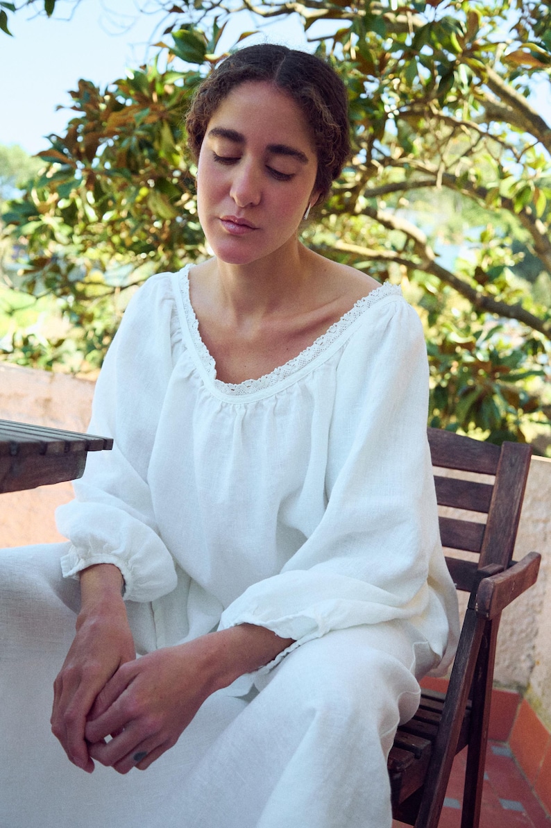 May include: A person wearing a white linen top and pants. The top has a scoop neckline with lace trim and long sleeves. The person is seated on a wooden chair, with a natural background of trees and foliage.