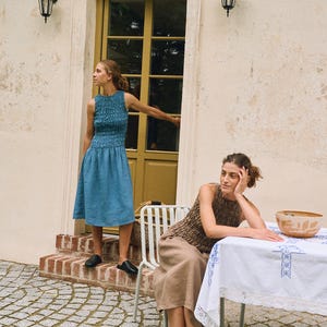 May include: Two women in dresses pose outdoors. One wears a blue sleeveless dress with a textured bodice and a flared skirt. The other wears a brown textured top and a beige skirt. They are near a table with a white tablecloth.