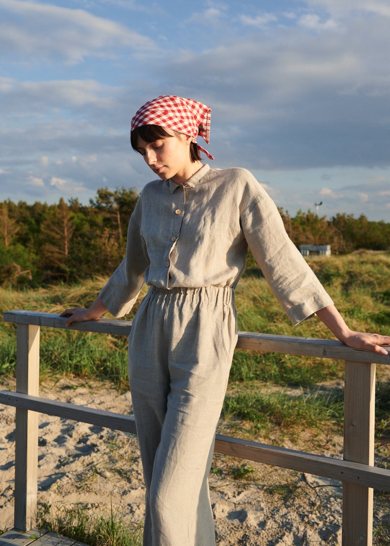 May include: A woman wearing a red and white checkered bandana, a light beige linen jumpsuit, and standing on a wooden railing. The background is a grassy field with trees and a blue sky.