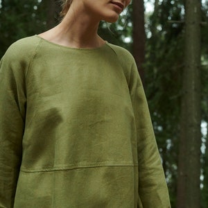 Peut inclure: Une femme portant une robe en lin verte &agrave; la coupe ample et &agrave; l'encolure ronde. La robe a une couture &agrave; la taille et des manches longues avec des poignets retrouss&eacute;s. La femme est debout dans un environnement forestier.