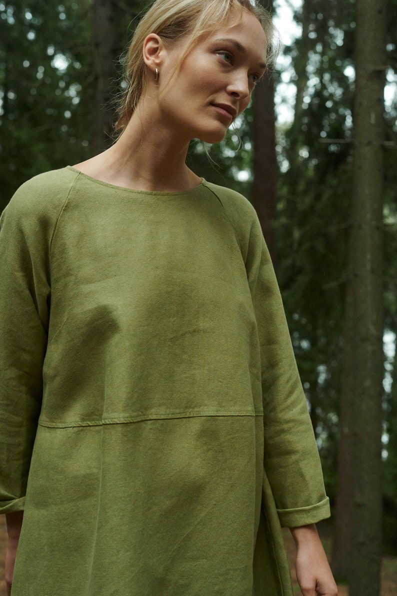 Peut inclure: Une femme portant une robe en lin verte &agrave; la coupe ample et &agrave; l'encolure ronde. La robe a une couture &agrave; la taille et des manches longues avec des poignets retrouss&eacute;s. La femme est debout dans un environnement forestier.