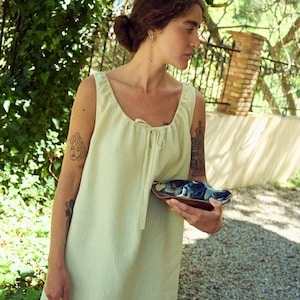 May include: A light green sleeveless dress with a square neckline and tie detail. The dress has a subtle checkered pattern. A small, decorative ceramic dish with blue and brown glaze is held in the hand.