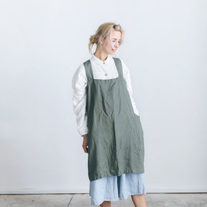May include: A woman wearing a green linen apron over a white shirt and a light blue skirt. She is standing on a concrete floor.