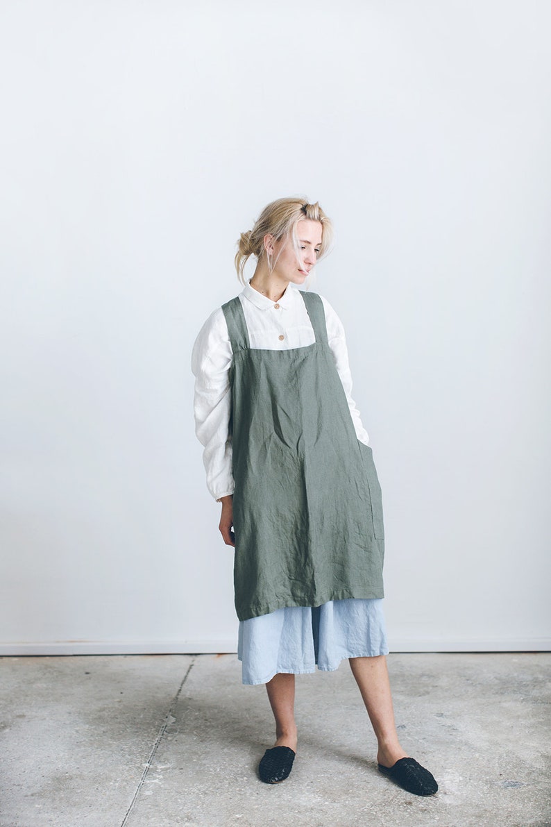 May include: A woman wearing a green linen apron over a white shirt and a light blue skirt. She is standing on a concrete floor.