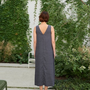 Puede incluir: Una mujer con un vestido de lino gris sin mangas de pie frente a una pared blanca cubierta de hiedra verde.