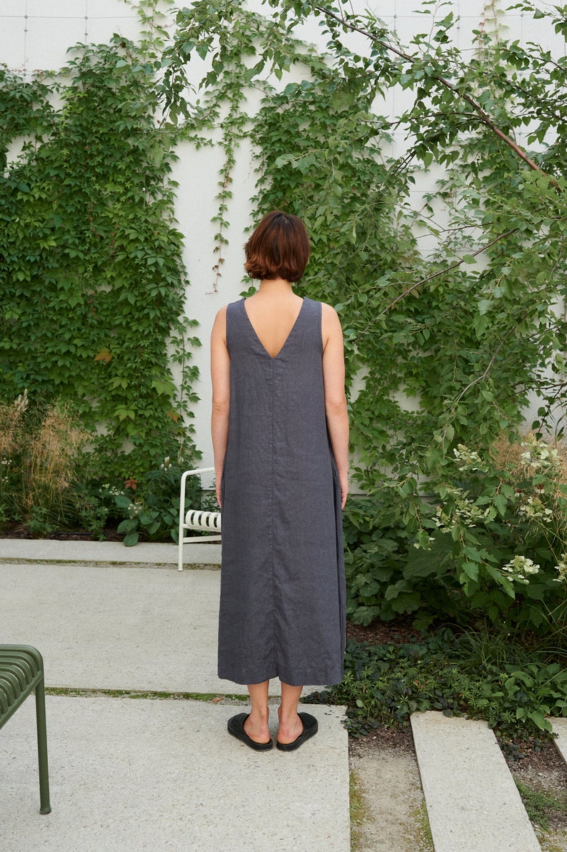 Puede incluir: Una mujer con un vestido de lino gris sin mangas de pie frente a una pared blanca cubierta de hiedra verde.