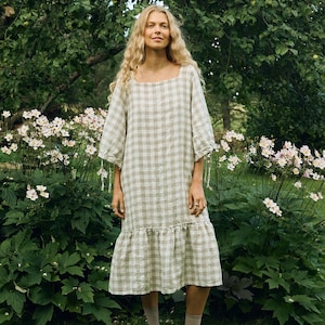 May include: A woman wearing a light green and white checkered linen dress with a square neckline and ruffled hem. The dress has three-quarter sleeves with tie details. She is wearing tan shoes and socks, standing in a garden with white flowers and green foliage.