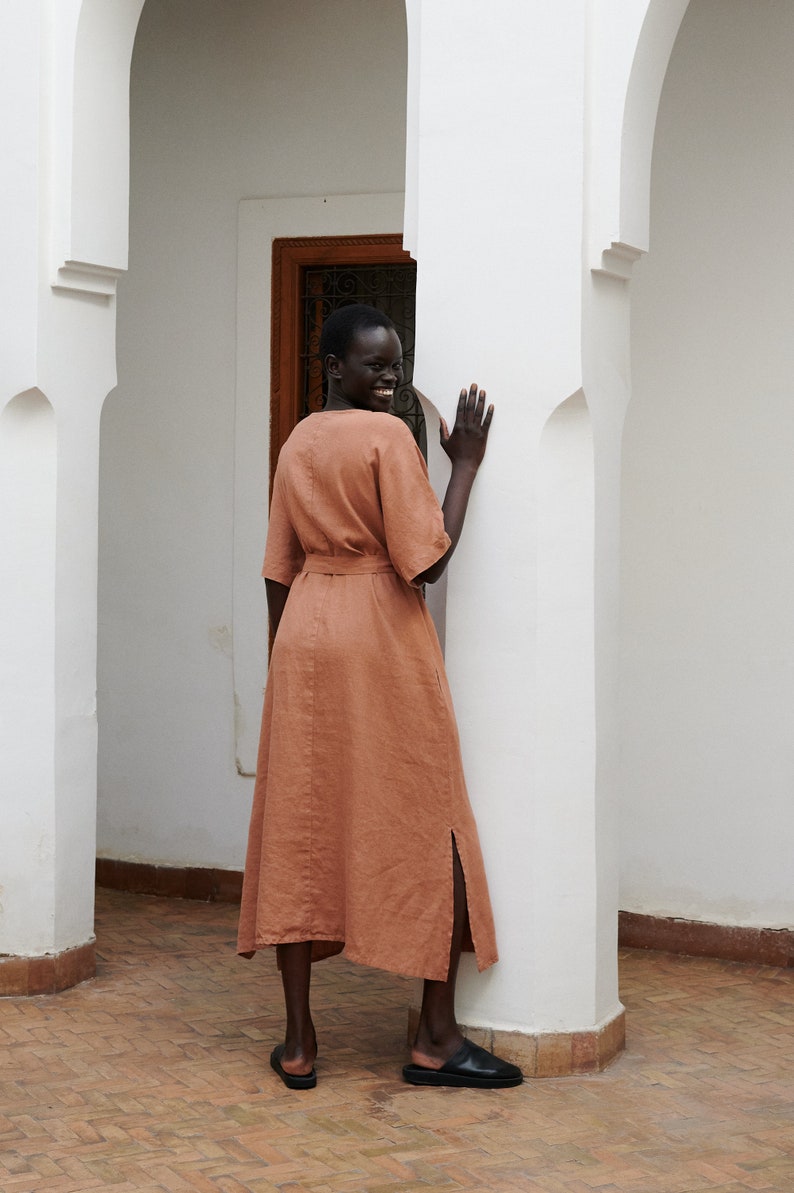 Puede incluir: Una mujer lleva un vestido de lino terracota con cintur&oacute;n y una abertura lateral. Est&aacute; de pie en una puerta arqueada blanca y sonr&iacute;e.