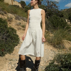 May include: A white sleeveless dress with a smocked bodice and a flowing skirt. The dress is worn with black knee-high boots.