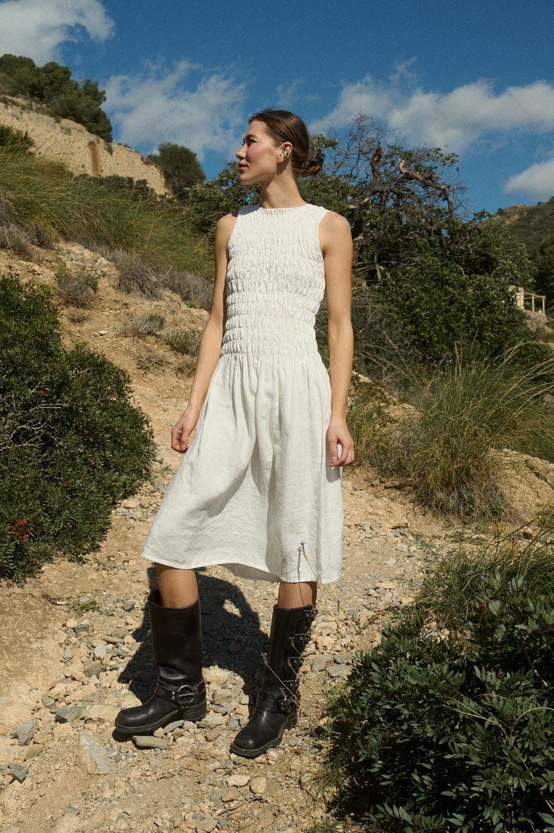 May include: A white sleeveless dress with a smocked bodice and a flowing skirt. The dress is worn with black knee-high boots.