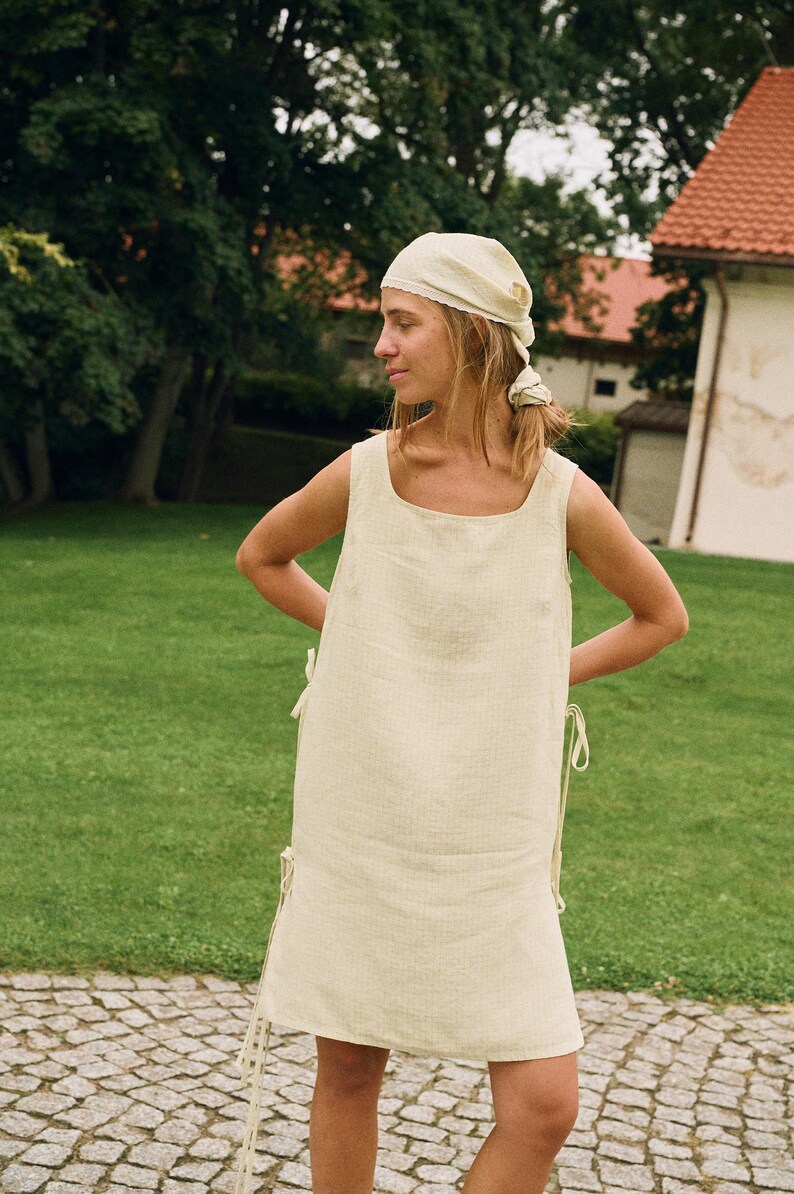 May include: A light yellow linen dress with a square neckline and tie details on the sides. The dress is sleeveless and worn with a matching headscarf. The background features a green lawn and a building with a red roof.