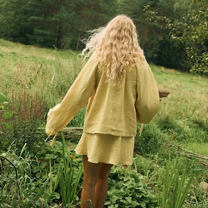 Pode incluir: Uma pessoa com um conjunto de linho amarelo, incluindo uma parte superior de manga comprida e uma saia, est&aacute; num jardim verde. A cor do traje harmoniza com o ambiente natural. A pessoa tem cabelo loiro comprido e ondulado. A cena evoca beleza natural e simplicidade.