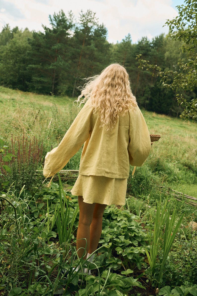 Pode incluir: Uma pessoa com um conjunto de linho amarelo, incluindo uma parte superior de manga comprida e uma saia, est&aacute; num jardim verde. A cor do traje harmoniza com o ambiente natural. A pessoa tem cabelo loiro comprido e ondulado. A cena evoca beleza natural e simplicidade.