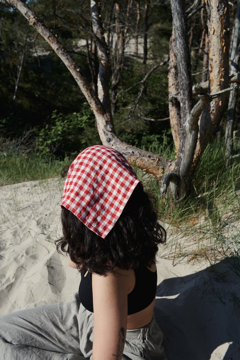 May include: A person wearing a red and white checkered bandana on their head, sitting on a sandy beach. They are wearing a black tank top and gray pants.