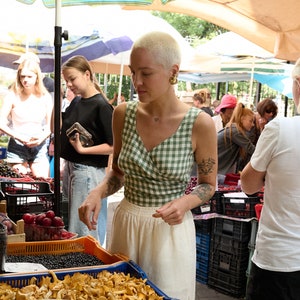 May include: A woman wearing a green and white gingham tank top and white trousers browses the produce at an outdoor market. She is looking at a selection of mushrooms and cucumbers.