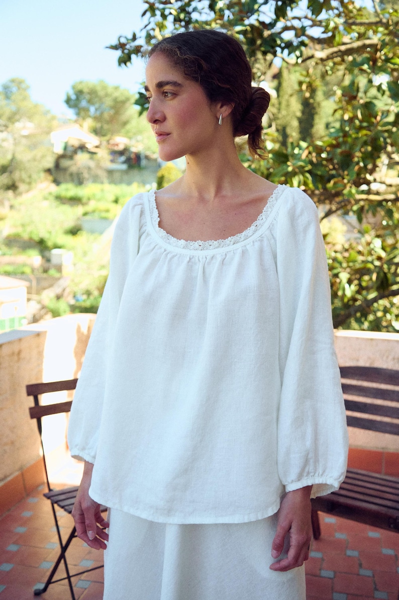 May include: White linen blouse with a square neckline and lace trim. The long sleeves gather at the wrist. The blouse is part of a two-piece set. The model is standing outside on a sunny day.