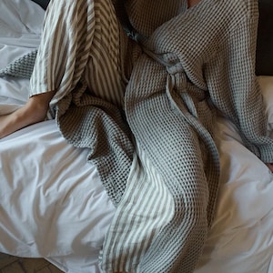 May include: A gray waffle weave robe with a striped linen pants. The robe is tied at the waist and the person is sitting on a white bed.