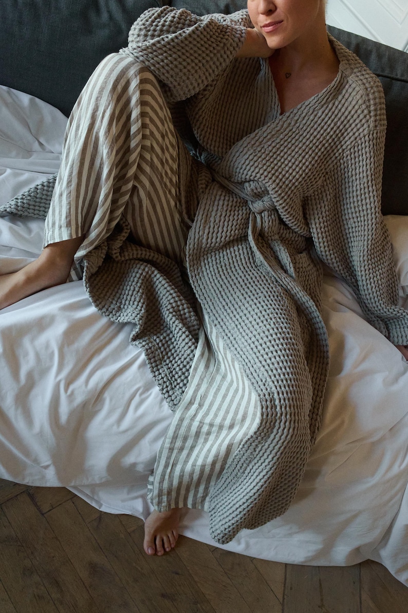May include: A gray waffle weave robe with a striped linen pants. The robe is tied at the waist and the person is sitting on a white bed.
