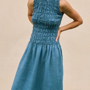 May include: A sleeveless, knee-length dress in a muted teal color. The bodice features a smocked design, while the skirt has a flowing, gathered style. The dress is made of a soft, lightweight fabric, perfect for warm weather.