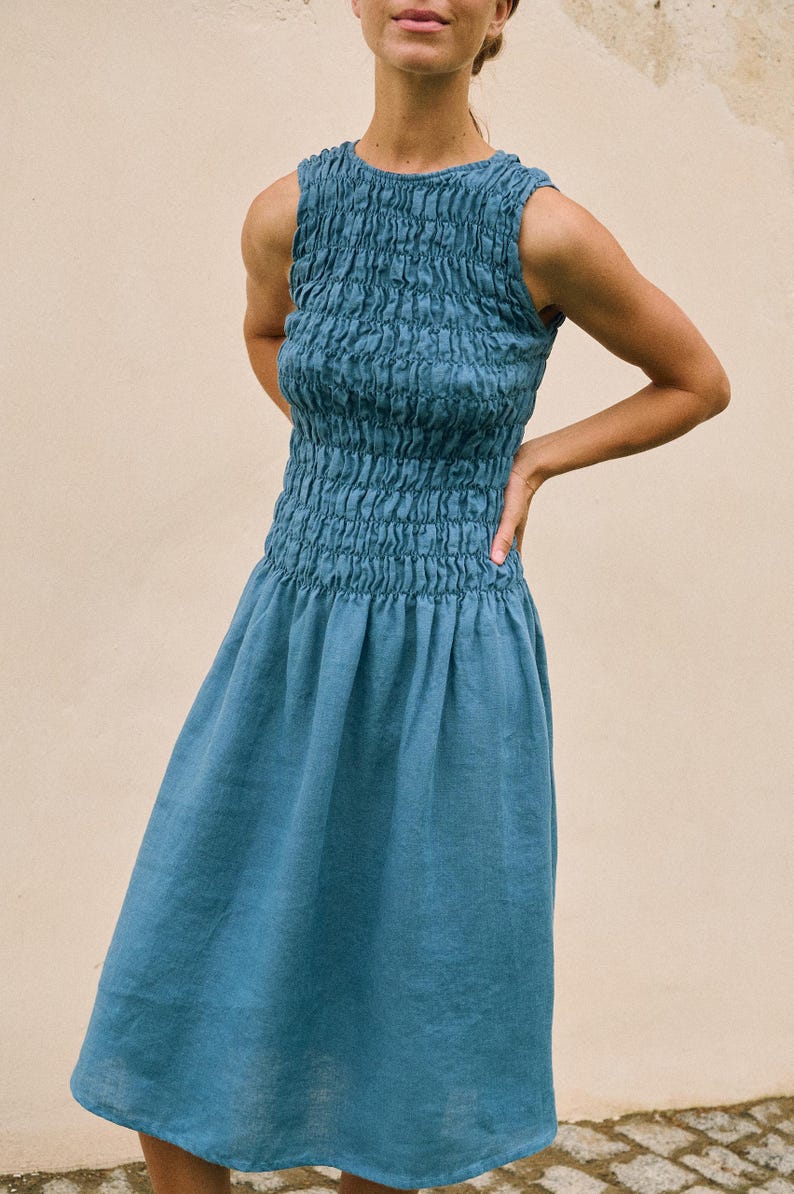 May include: A sleeveless, knee-length dress in a muted teal color. The bodice features a smocked design, while the skirt has a flowing, gathered style. The dress is made of a soft, lightweight fabric, perfect for warm weather.