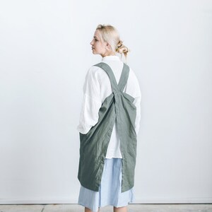 May include: A woman wearing a long, sage green linen apron with a criss-cross back strap. The apron is tied at the waist and has a large pocket on the front. She is wearing a white shirt and a light blue skirt.