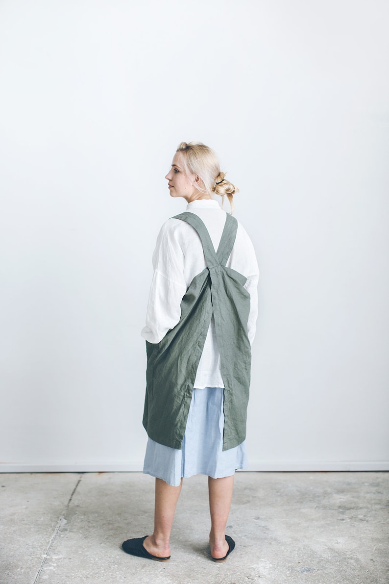 May include: A woman wearing a long, sage green linen apron with a criss-cross back strap. The apron is tied at the waist and has a large pocket on the front. She is wearing a white shirt and a light blue skirt.