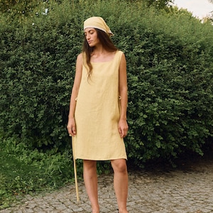 May include: A sleeveless, pale yellow linen dress with a square neckline and side ties. The dress is knee-length and worn with a matching headscarf and black flats. The background features lush green foliage and a stone path.