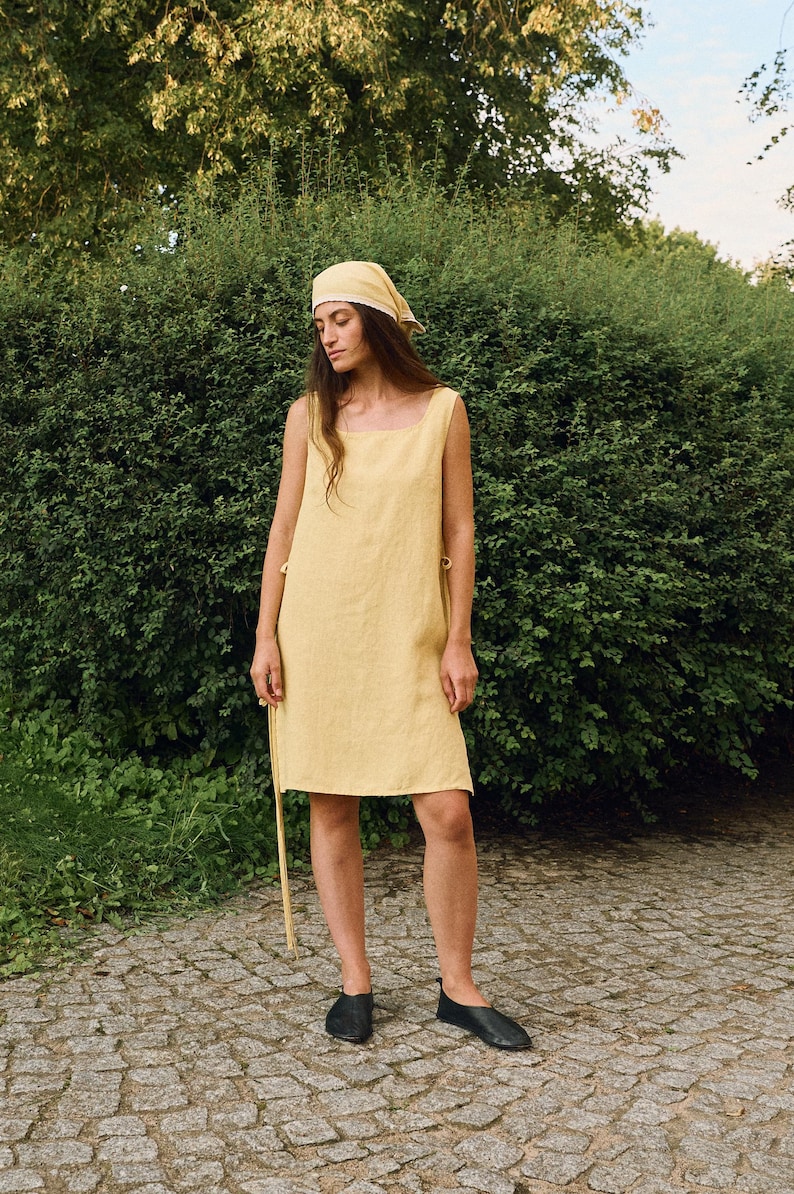 May include: A sleeveless, pale yellow linen dress with a square neckline and side ties. The dress is knee-length and worn with a matching headscarf and black flats. The background features lush green foliage and a stone path.