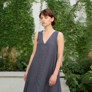 Puede incluir: Una mujer lleva un vestido de lino sin mangas de color gris oscuro con escote en V. Est&aacute; de pie frente a una pared cubierta de hiedra verde.