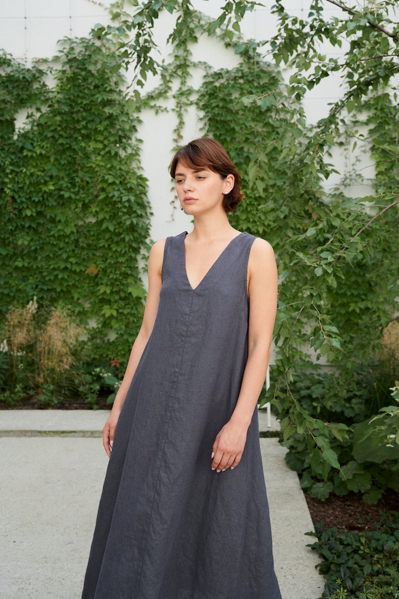 Puede incluir: Una mujer lleva un vestido de lino sin mangas de color gris oscuro con escote en V. Est&aacute; de pie frente a una pared cubierta de hiedra verde.