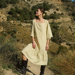 May include: A woman wearing a light yellow linen dress with a square neckline and a ruffled hem. She is standing in a field of tall grass and looking up at the sky.