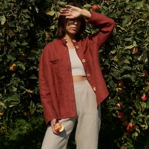 May include: A woman wearing a red linen shirt with a white tank top underneath and beige linen pants. She is standing in front of an apple tree and holding a red apple in her hand.