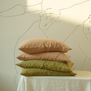 Puede incluir: Una pila de cuatro almohadas de lino en tonos melocotón y verde oliva. Las almohadas están colocadas sobre un mantel blanco. El fondo presenta una pared con un diseño en relieve claro.