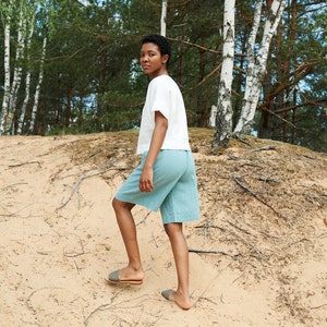 May include: A person wearing a white linen top and light blue linen shorts walks on a sandy path in a birch tree forest.