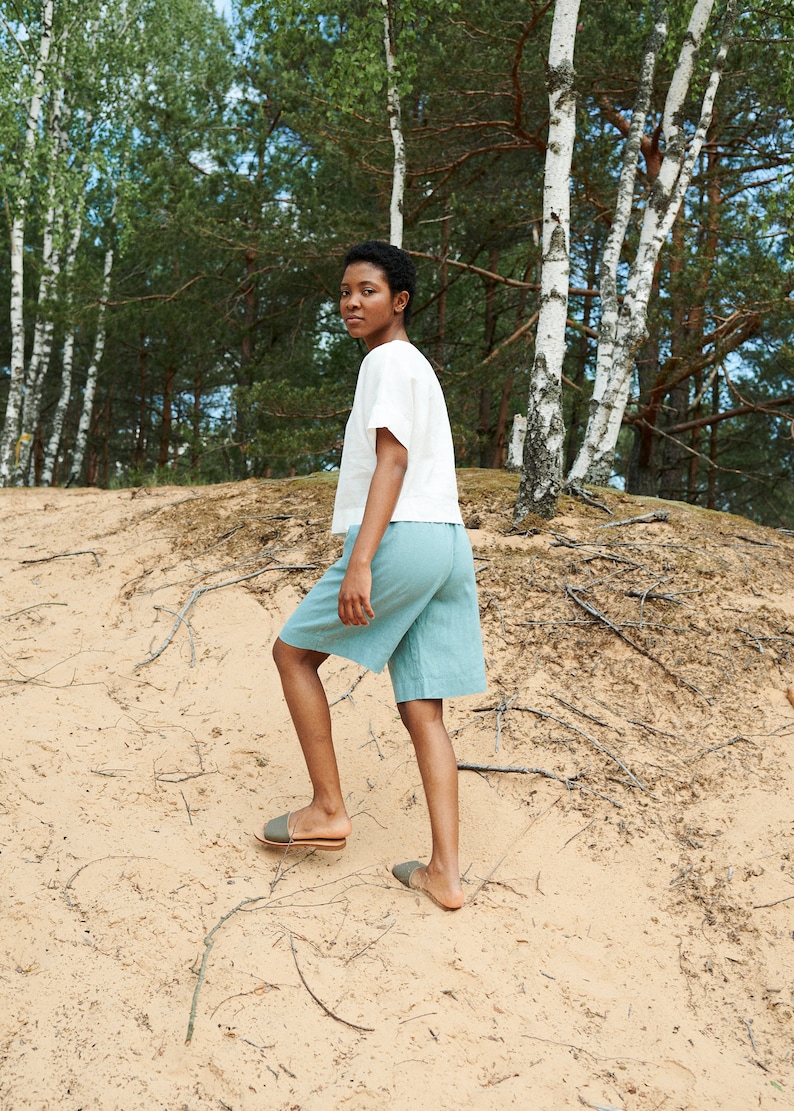 May include: A person wearing a white linen top and light blue linen shorts walks on a sandy path in a birch tree forest.