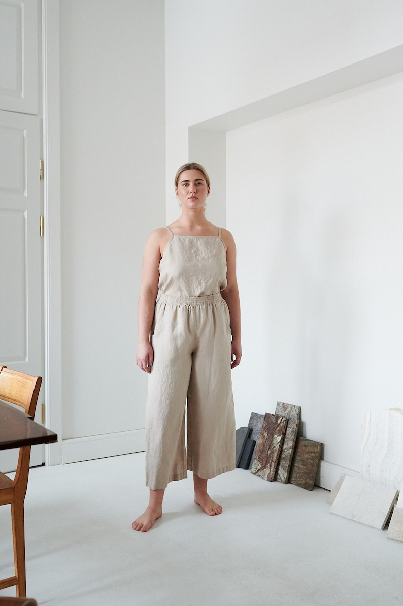 May include: A woman wearing a beige linen jumpsuit with a square neckline and wide legs. She is standing in a white room with a wooden table and some decorative tiles on the floor.