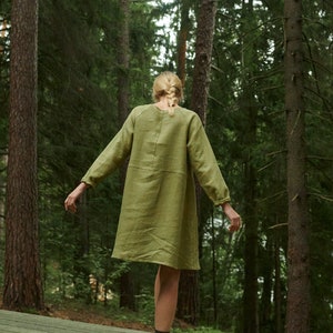 Peut inclure: Une femme portant une robe en lin vert et des chaussettes noires se tient sur une plateforme en bois dans une for&ecirc;t. Elle regarde loin de l'appareil photo.