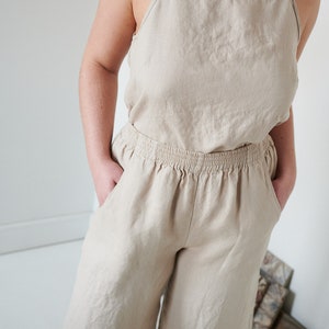 May include: A light beige linen jumpsuit with a sleeveless top and wide-leg pants. The pants have an elastic waistband and pockets.