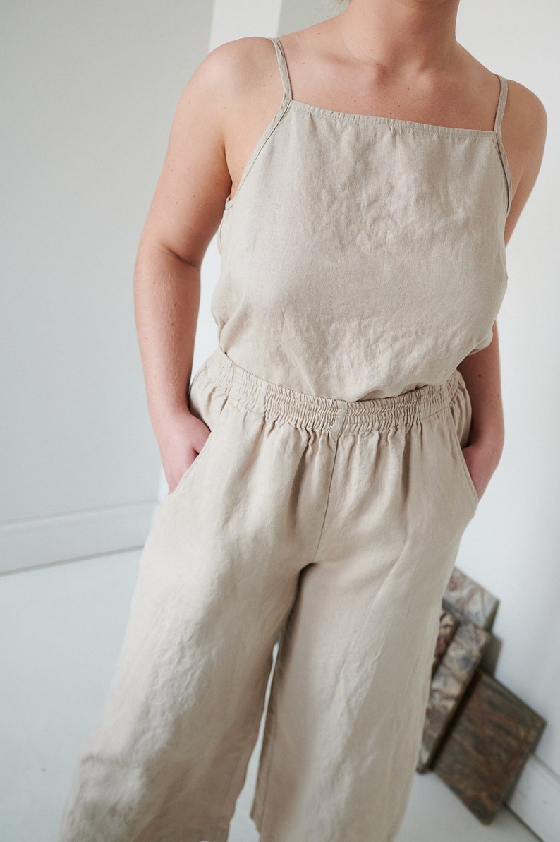 May include: A light beige linen jumpsuit with a sleeveless top and wide-leg pants. The pants have an elastic waistband and pockets.